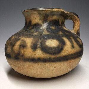 May include: A small, round, ceramic pitcher with a handle. The pitcher is tan with dark brown abstract designs around the body. The rim and handle are also dark brown. The pitcher has a rustic, handmade appearance.
