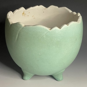 May include: A light green ceramic planter with a unique eggshell design. The planter has a scalloped rim and three small feet. The interior is off-white, and the exterior has a crackled texture. A decorative piece for plants.