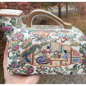 May include: A white porcelain pitcher with a gold handle and spout. The pitcher is decorated with colourful floral and figural scenes. The handle is a curved, gold-coloured metal. The overall design is ornate and detailed.