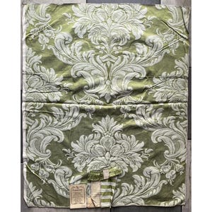 May include: A rectangular piece of sage green fabric featuring a repeating white damask pattern with intricate floral and scrollwork motifs. A small rectangular paper tag with printed text is attached to the bottom edge of the textile.