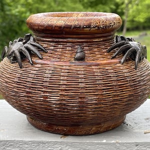 May include: A decorative, handcrafted basket-style vase with a wide, open top. The vase is made of woven brown material and features dark crab-shaped embellishments around the rim. A small, dark snail-shaped accent is centered on the front.