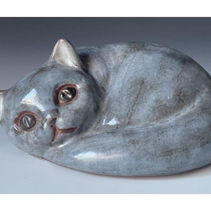 Puede incluir: Figura de gato de cerámica en posición enroscada. El gato es de color gris azulado apagado con veteado gris más oscuro. Tiene ojos marrones y boca rojiza. Las orejas y la punta de la cola son blancas.