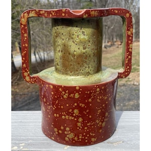 May include: A handcrafted ceramic vase with a unique design. The vase features a cylindrical body in a deep red hue, speckled with gold-coloured spots. It has a green and gold interior and a rectangular handle. The vase is a decorative piece.