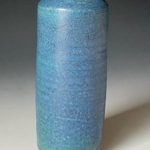 May include: A tall, cylindrical ceramic vase with a flared rim, featuring a textured, mottled blue glaze that transitions to a lighter turquoise hue near the base, revealing a thin band of unglazed reddish-brown clay at the bottom edge.