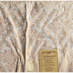 May include: Close-up of a cream-colored fabric with a repeating floral pattern in a light gold color. The fabric is folded and has a tag attached with the name "Scalamandré" and the number "92209-1".