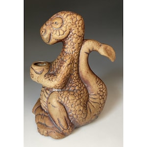 May include: A tan ceramic figurine of a seated dinosaur with a textured, scaly surface. The creature has a long, curved tail and holds a small, circular bowl-shaped opening in its front paws. The design features dark brown detailing in the crevices.