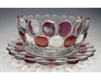 Puede incluir: Un cuenco de vidrio transparente y un platillo a juego con un patrón en relieve de círculos rosas y blancos. El cuenco y el platillo están hechos de vidrio y tienen un aspecto vintage.