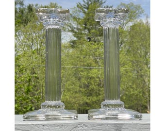 May include: Two clear glass candle holders shaped like columns with fluted details. The candle holders are on a white surface with a green background.