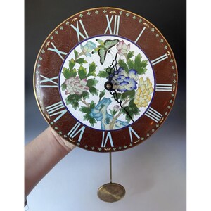 May include: Round wall clock with a brown face and Roman numerals. The clock features a floral design with butterflies and a pendulum. The clock's face is brown with a gold trim.