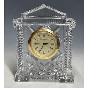 Puede incluir: Un reloj de cristal de Waterford con una esfera y agujas doradas. El reloj está en una caja de cristal transparente con un diseño decorativo.