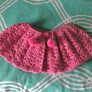 May include: A pink crocheted cape with a button closure and two pom-poms.