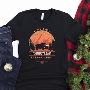 May include: Black t-shirt with a graphic of a buffalo wearing a Santa hat in front of a mountain scene with Christmas lights. The text "This is my Christmas Pajama Shirt" is printed on the shirt.