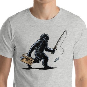 May include: A heather grey t-shirt featuring a black and white illustration of a Bigfoot holding a fishing rod and a tackle box.
