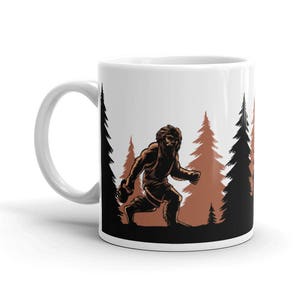 May include: White ceramic coffee mug with a brown silhouette of a Bigfoot walking through a forest of brown trees.
