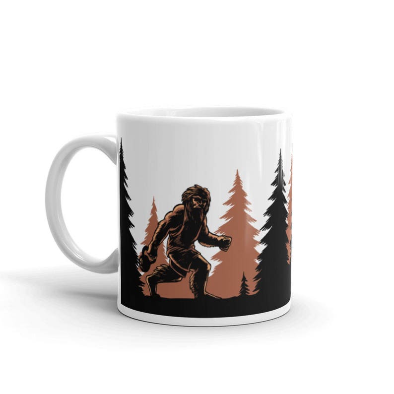 May include: White ceramic coffee mug with a brown silhouette of a Bigfoot walking through a forest of brown trees.