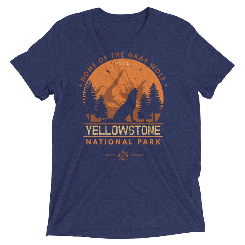May include: A navy blue t-shirt with an orange graphic of a wolf howling at the moon in a forest setting. The graphic includes the text "Home of the Gray Wolf", "Yellowstone National Park", and "1872".