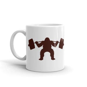 May include: White ceramic coffee mug with a brown silhouette of a gorilla lifting a barbell. The gorilla is squatting with its arms extended, holding the barbell.