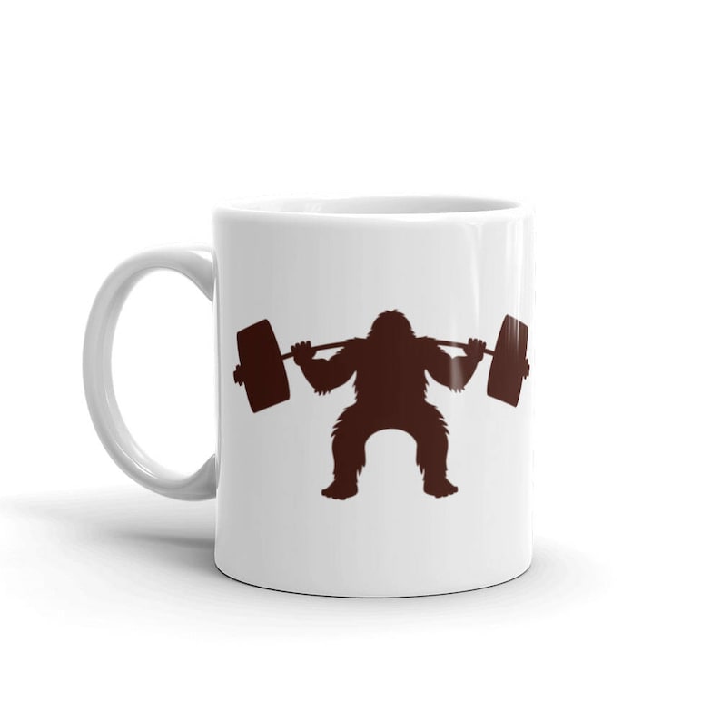May include: White ceramic coffee mug with a brown silhouette of a gorilla lifting a barbell. The gorilla is squatting with its arms extended, holding the barbell.