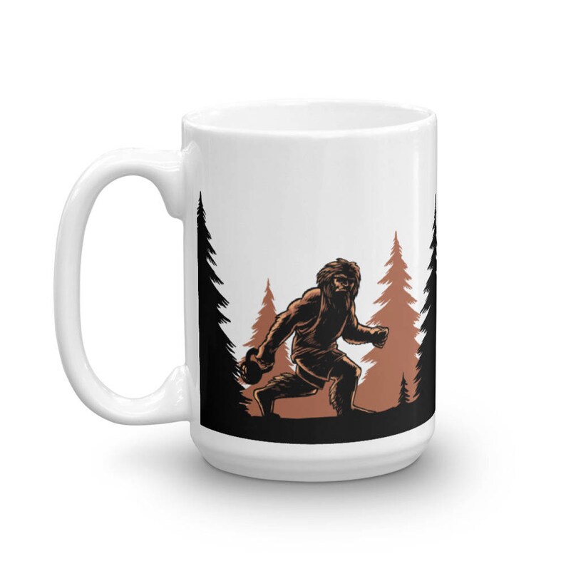 May include: White ceramic mug with a black and brown illustration of a Bigfoot figure walking through a forest. The Bigfoot is holding a ball in its hand.
