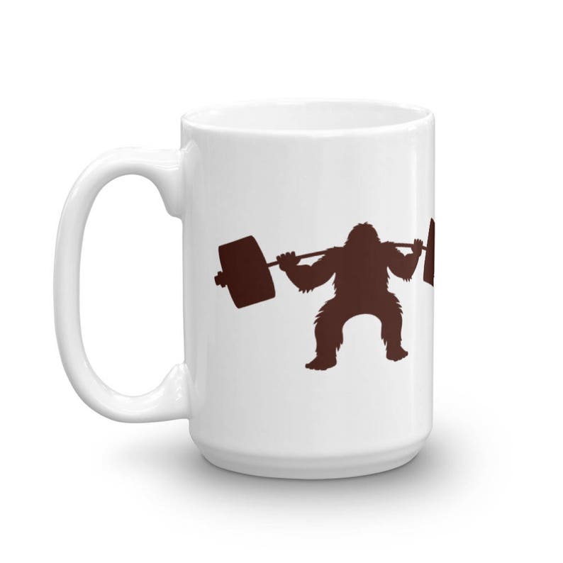 May include: White ceramic coffee mug with a brown silhouette of a creature lifting a barbell. The creature has a hairy body and is squatting.