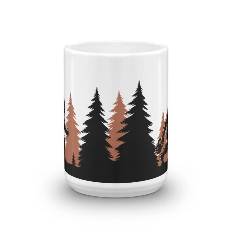 May include: White ceramic mug with a black and brown silhouette design of trees. The trees are in a forest setting with a brown silhouette of a creature in the background.