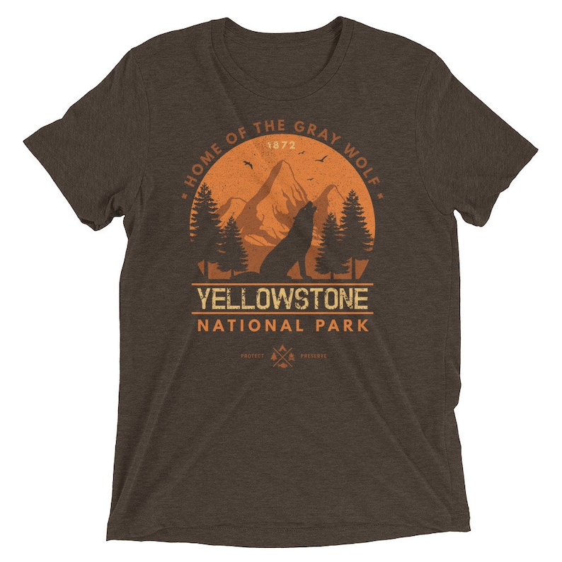May include: Brown t-shirt with a vintage-style graphic of a wolf howling at the moon in front of a mountain range. The graphic also includes the text "Home of the Gray Wolf", "Yellowstone National Park", and "1872".