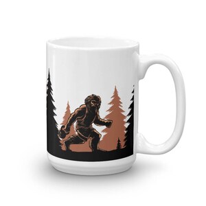 May include: White ceramic mug with a black and brown illustration of a Bigfoot walking through a forest. The Bigfoot is in the center of the image, with trees on either side.