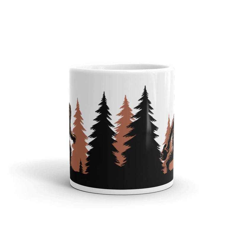 May include: White ceramic coffee mug with a black and brown silhouette design of a Bigfoot walking through a forest of trees.