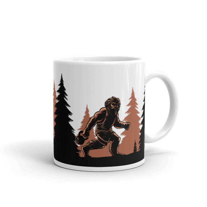 May include: White ceramic mug with a brown silhouette of a Bigfoot figure holding a dumbbell in a forest setting. The Bigfoot is walking towards the right side of the mug. The trees are brown and the background is white.