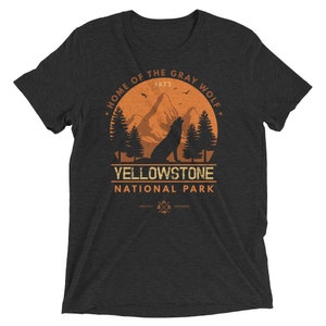 May include: A dark gray t-shirt with a vintage-style graphic of a wolf howling at the moon in a forest setting. The graphic includes the text "Home of the Gray Wolf", "Yellowstone National Park", and "1872".