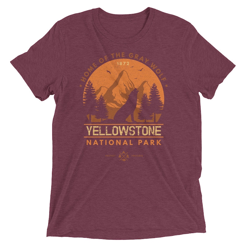 May include: A maroon t-shirt with a vintage-style graphic of a wolf howling at the moon in front of a mountain range. The graphic includes the text "Home of the Gray Wolf", "Yellowstone National Park", and "1872".