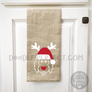 Pu&ograve; includere: Un asciugamano da cucina beige con un doodle bianco di un cane che indossa un cappello rosso di Babbo Natale e corna di renna. Il testo "Doodleperfect.com" &egrave; stampato sull'asciugamano.