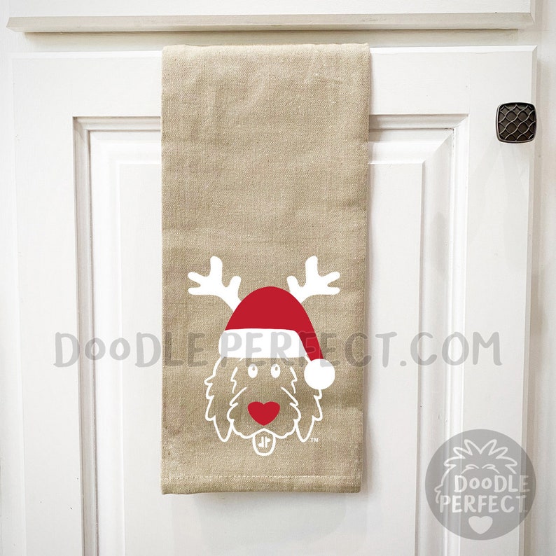 Pu&ograve; includere: Un asciugamano da cucina beige con un doodle bianco di un cane che indossa un cappello rosso di Babbo Natale e corna di renna. Il testo "Doodleperfect.com" &egrave; stampato sull'asciugamano.