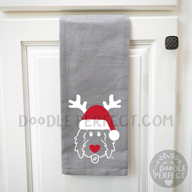 Pu&ograve; includere: Un asciugamano da cucina grigio con un doodle bianco di un cane che indossa un cappello rosso di Babbo Natale e corna di renna. Il testo "Doodle Perfect.com" &egrave; stampato sull'asciugamano.