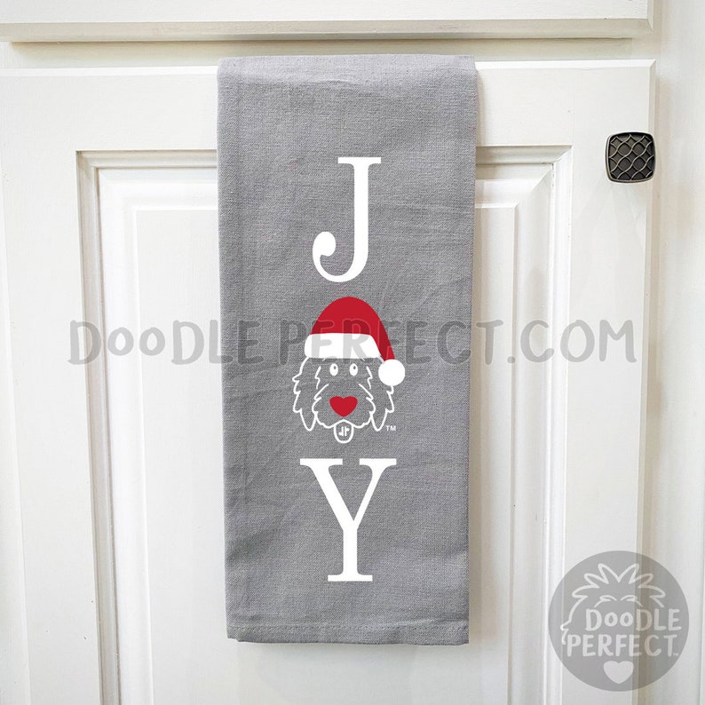 Pu&ograve; includere: Un asciugamano da cucina grigio con la parola "JOY" stampata in bianco. La "O" &egrave; sostituita da un doodle di un cane che indossa un cappello da Babbo Natale rosso.
