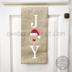 Pu&ograve; includere: Un asciugamano da cucina beige con la parola "JOY" stampata in bianco. La "O" &egrave; sostituita da un doodle di un cane che indossa un cappello da Babbo Natale rosso.