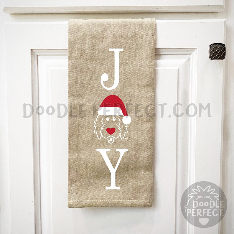 Pu&ograve; includere: Un asciugamano da cucina beige con la parola "JOY" stampata in bianco. La "O" &egrave; sostituita da un doodle di un cane che indossa un cappello da Babbo Natale rosso.