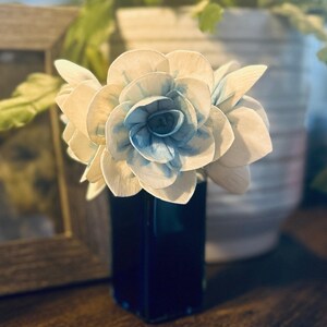 May include: A bouquet of white and blue paper flowers in a dark blue vase. The flowers have a delicate, layered design.