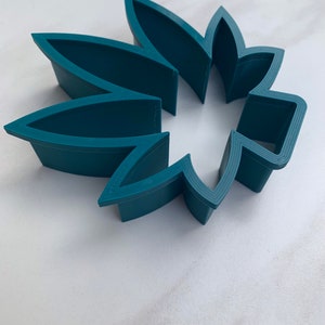 May include: A teal-colored, flower-shaped cookie cutter. The cutter is made of plastic and has a series of petal-shaped cutouts. The cutter is designed to cut out flower shapes from dough or fondant for baking or crafting projects.