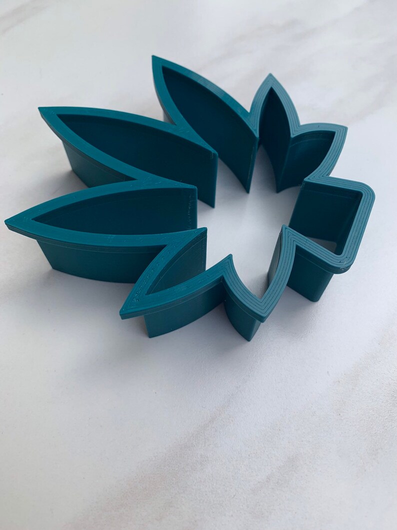 May include: A teal-colored, flower-shaped cookie cutter. The cutter is made of plastic and has a series of petal-shaped cutouts. The cutter is designed to cut out flower shapes from dough or fondant for baking or crafting projects.