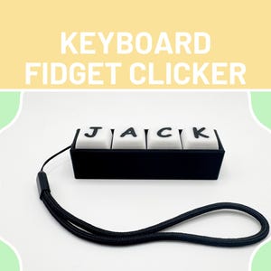 Peut inclure: Un clicker de clavier noir avec le nom "JACK" sur des touches blanches. Le clicker est muni d'une dragonne noire. Le texte "KEYBOARD FIDGET CLICKER" est en haut de l'image.