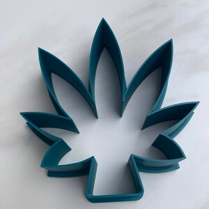 May include: A teal-colored, leaf-shaped cookie cutter. The cutter is made of plastic and has a detailed edge. The design features seven distinct points, creating a realistic leaf shape. The cutter is designed for baking and crafting.