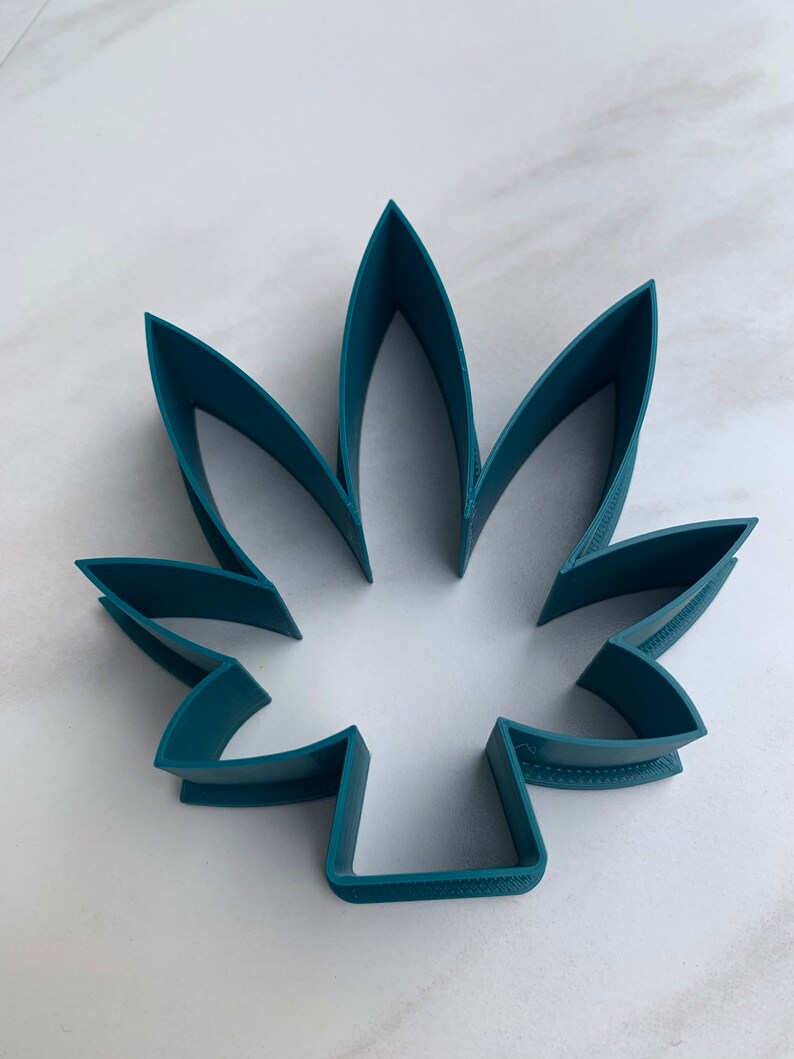 May include: A teal-colored, leaf-shaped cookie cutter. The cutter is made of plastic and has a detailed edge. The design features seven distinct points, creating a realistic leaf shape. The cutter is designed for baking and crafting.