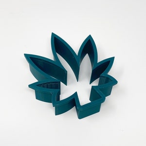 May include: A teal-colored, leaf-shaped cookie cutter. The cutter is made of plastic and has a detailed design, with the leaf's veins clearly visible. The cutter is designed to cut out the shape of a cannabis leaf.
