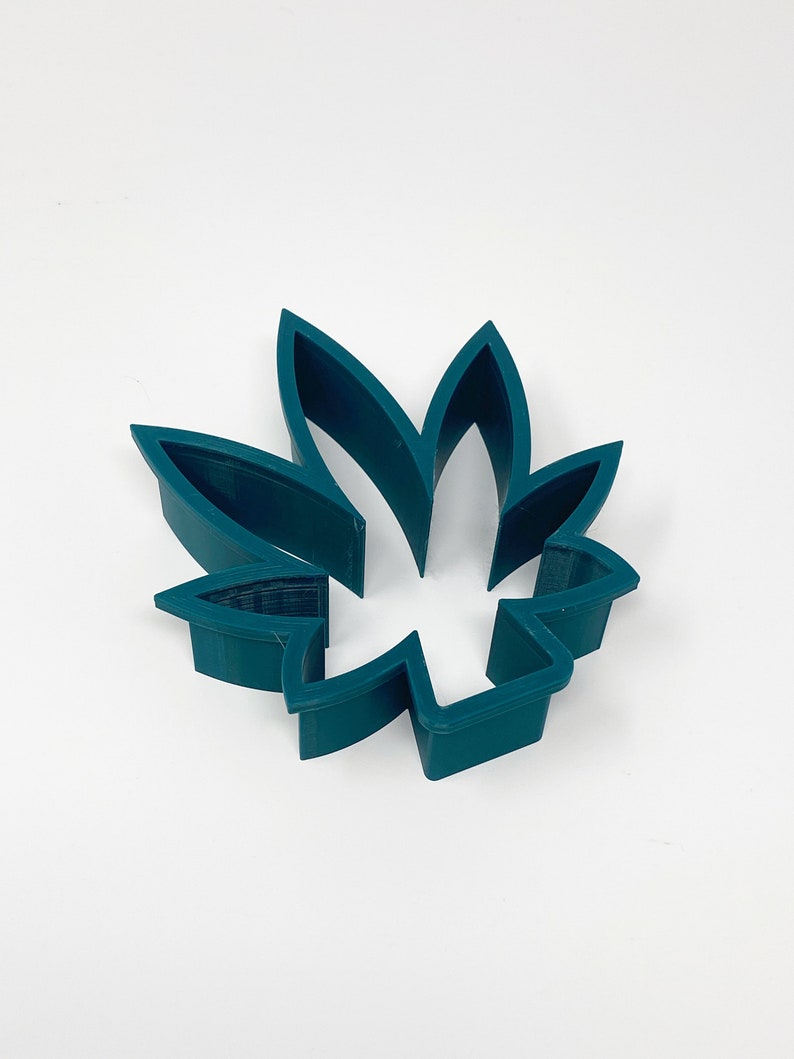 May include: A teal-colored, leaf-shaped cookie cutter. The cutter is made of plastic and has a detailed design, with the leaf's veins clearly visible. The cutter is designed to cut out the shape of a cannabis leaf.