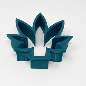 May include: A teal-colored, five-petal flower-shaped cookie cutter. The cutter is made of plastic and has a simple, geometric design. The petals are pointed and the base is rectangular.
