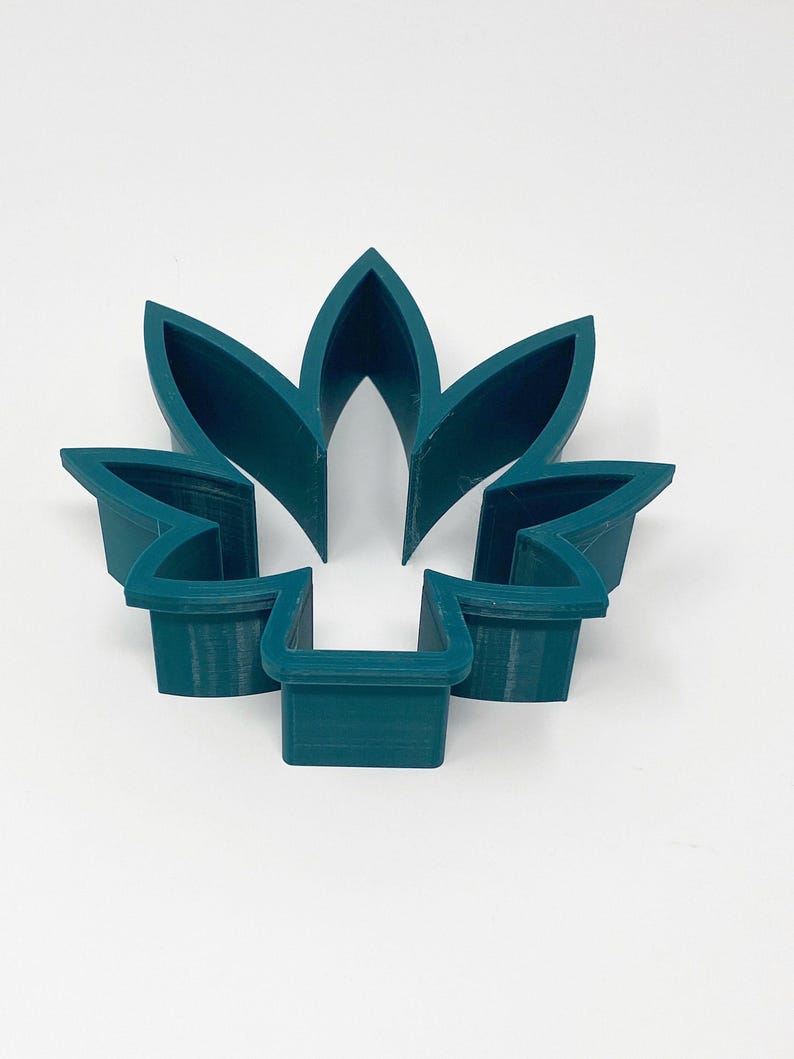 May include: A teal-colored, five-petal flower-shaped cookie cutter. The cutter is made of plastic and has a simple, geometric design. The petals are pointed and the base is rectangular.