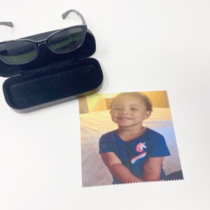 May include: Black sunglasses with a black case and a microfiber cloth with a smiling child wearing a blue shirt with a unicorn and rainbow graphic. The sunglasses have a sleek, modern design.