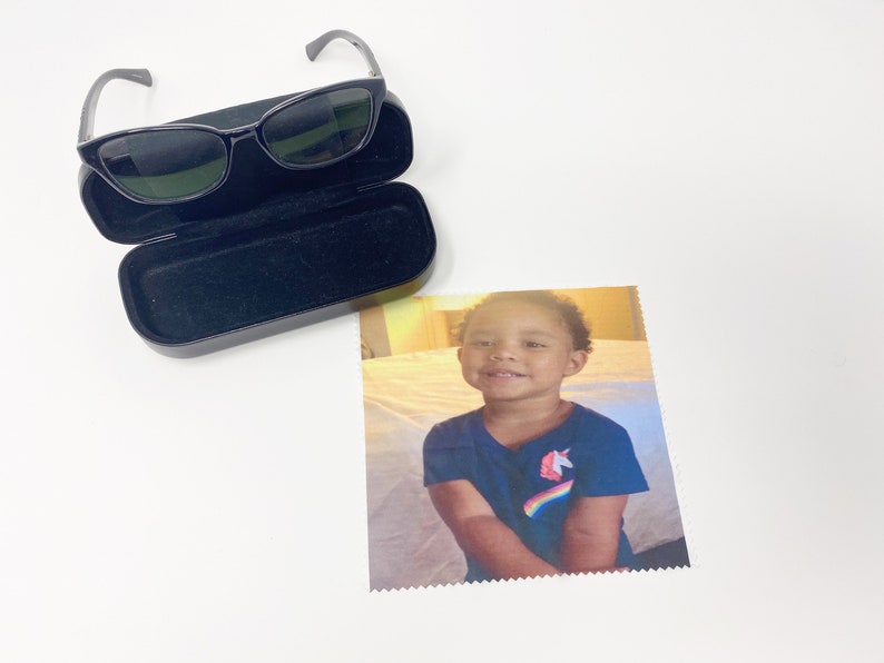 May include: Black sunglasses with a black case and a microfiber cloth with a smiling child wearing a blue shirt with a unicorn and rainbow graphic. The sunglasses have a sleek, modern design.