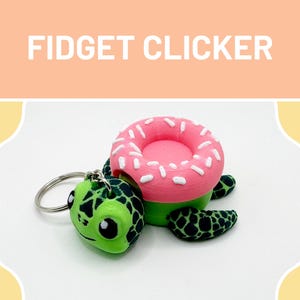 May include: A novelty fidget clicker featuring a turtle and a donut design. The turtle is green with black markings and a smiling face. The donut is pink with white sprinkles, set on a green base. The words "FIDGET CLICKER" are displayed above the objects.